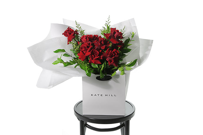 Classic and large red rose bouquet displaying 12 stems of red reflexed roses and seasonal green foliage. Valentines day or Romance gift bouquet presented in our Kate Hill Flower Bag. DIOR boasts a collection of short to medium-length red roses intertwined with seasonal foliage. Bouquet Bag sitting on a black bentwood chair.