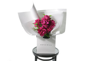 Best selling hot pink rose bouquet named Floyd featuring 6 stems of hot pink reflexed roses and green seasonal foliage. Gift bouquet presented in our Kate Hill Flower Bag. Floyd rose bouquet is sitting on a black bentwood chair.