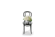 Wide image of FREDERICK hydrangea vase design to give size perspective. White contemporary ceramic ball vase displaying a dome of white hydrangeas only. Chic white hydrangea design is sitting on a black bentwood chair. Vase Design will be presented in a Kate Hill flower bag and be detailed with ribbon.