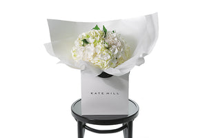 Small chic bouquet displaying a dome of white hydrangeas (no additional foliage). Bouquet presented in Kate Hill Flower Bag. Bouquet bag is sitting on a black bentwood chair.