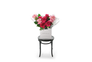Wide image of the GEORGIA rose bouquet. A large bouquet of mixed reflexed bright roses with lush green foliages. Beautifully wrapped in signature Kate Hill Flowers wrapping and placed into flower bag. Bouquet bag sitting on a black bentwood chair.