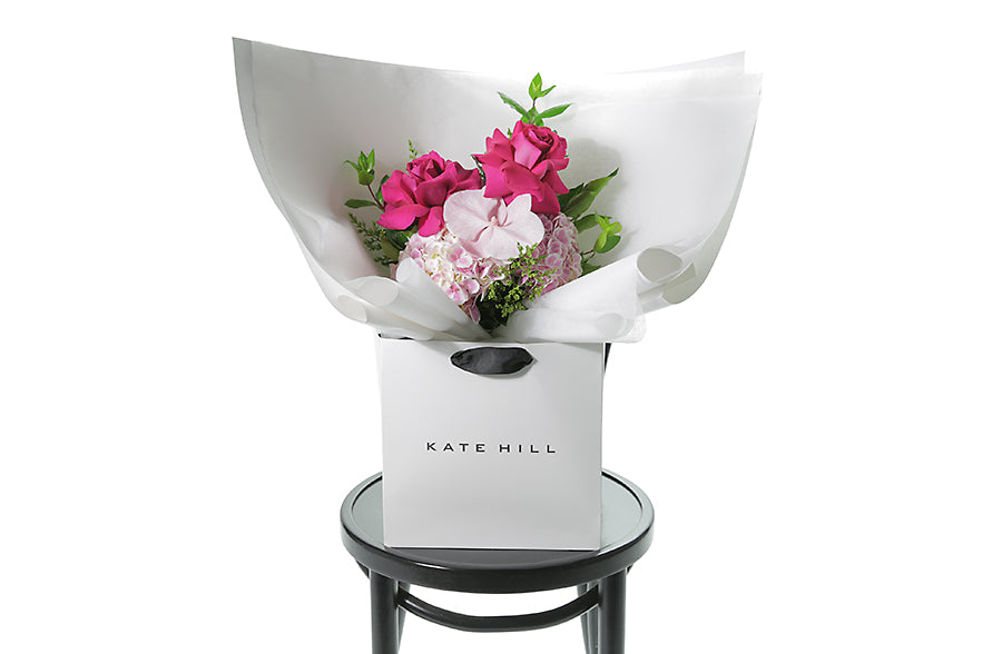 Small pastel pink and bright pink posy bouquet. Pink flowers and green seasonal foliage. Presented in Kate Hill Flower Bag. Image shows the Bouquet bag sitting on a black bentwood chair.