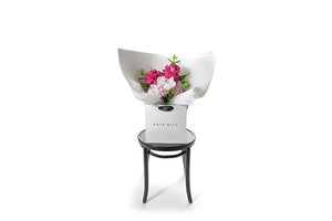Small pastel pink and bright pink posy bouquet. Pink flowers and green seasonal foliage. Presented in Kate Hill Flower Bag. Image shows the Bouquet bag sitting on a black bentwood chair. Wide image of bouquet bag so you can see true size of the bouquet.