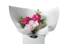 Small pastel pink and bright pink posy bouquet. Pink flowers and green seasonal foliage. Presented in Kate Hill Flower Bag. Image shows the Bouquet bag sitting on a black bentwood chair. Close up image of bouquet.