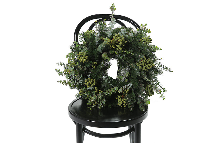 45cm-55cm Christmas Wreath displaying fragrant festive foliage including norwegian spruce foliage, spinning gum eucalyptus foliage and green berries. Wreath is presented with ribbons to hang christmas wreath to your front door or sit on table for a christmas table decor. Festive wreath is sitting on a black bentwood chair.