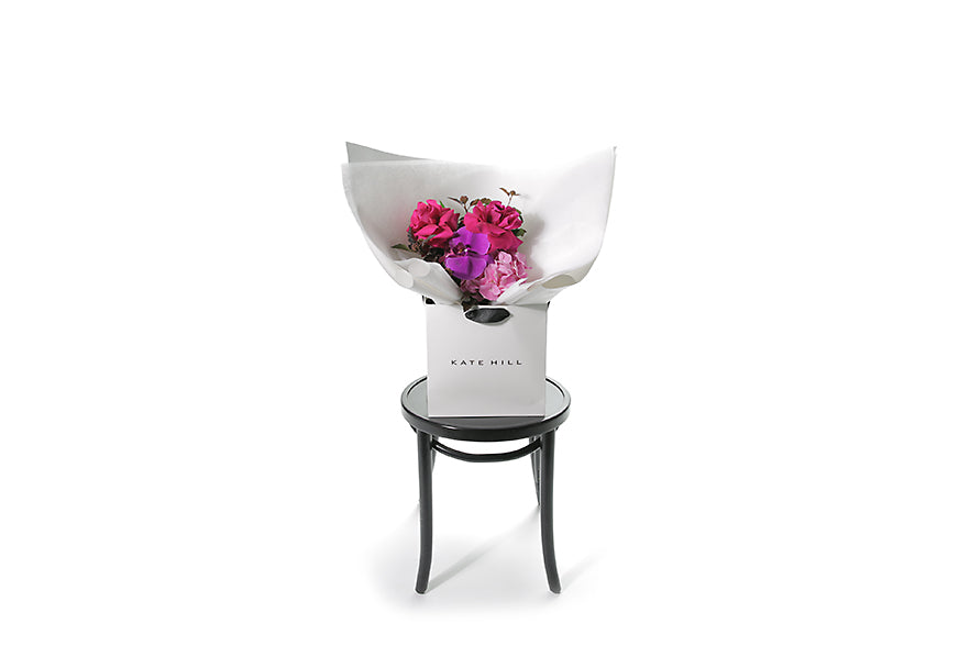 Wide image of the LENI flower bouquet. Petite in size and vibrant in colour. Small bright pink and magenta pink posy bouquet. Pink flowers and green seasonal foliage. Presented in Kate Hill Flower Bag. Bouquet bag is sitting on a black bentwood chair. 