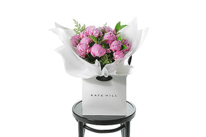 Beautiful pink peony rose bouquet displaying 15 stems of pink peony roses and green seasonal foliage. Bouquet of peony roses presented in white signature wrapping and placed into Kate Hill Flower Bag. Bouquet bag sitting on a black bentwood chair.
