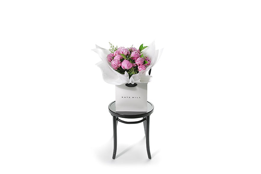 Wide image of the LUCIA peony rose flower bouquet so you can see the true size of the bouquet. Beautiful pink peony rose bouquet displaying 15 stems of pink peony roses and green seasonal foliage. Bouquet of peony roses presented in white signature wrapping and placed into Kate Hill Flower Bag. Bouquet bag sitting on a black bentwood chair.