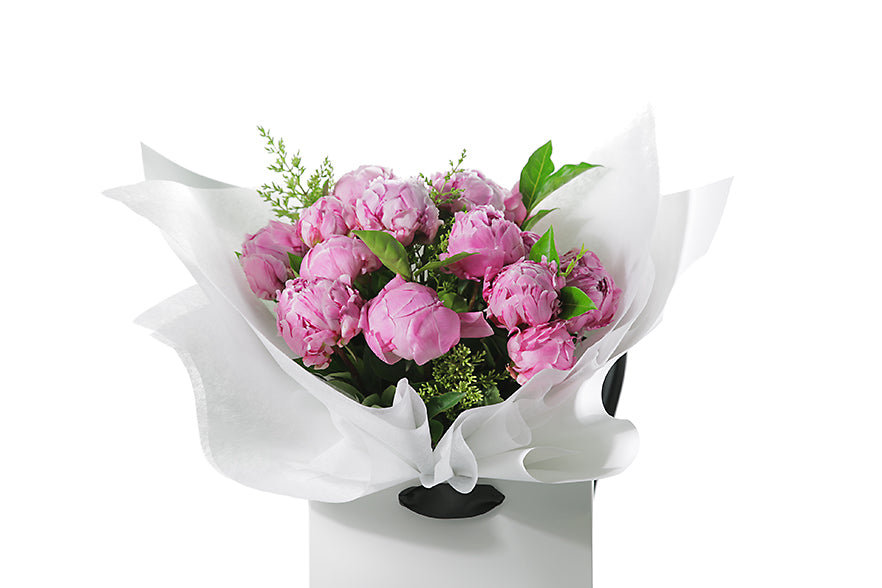 Close up image of the LUCIA peony rose bouquet. Beautiful pink peony rose bouquet displaying 15 stems of pink peony roses and green seasonal foliage. Bouquet of peony roses presented in white signature wrapping and placed into Kate Hill Flower Bag. Bouquet bag sitting on a black bentwood chair.