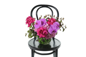 Ball Vase leaf lined with monsteria leaves, displaying bright pink, blush pink and magenta pink flowers and seasonal foliage. Vase included in gift. MAYA ball vase design sitting on a black bentwood chair.