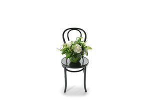 Wide image of the NOEL christmas vase design. NOEL festive vase design is a classic vase design that displays a balanced mix of white seasonal festive flowers and green festive foliages. A ball vase lined with green monsteria leaves, displaying festive foliages, berries, christmas lilies and seasonal white and green flower. Ball vase sitting on a black bentwood chair.
