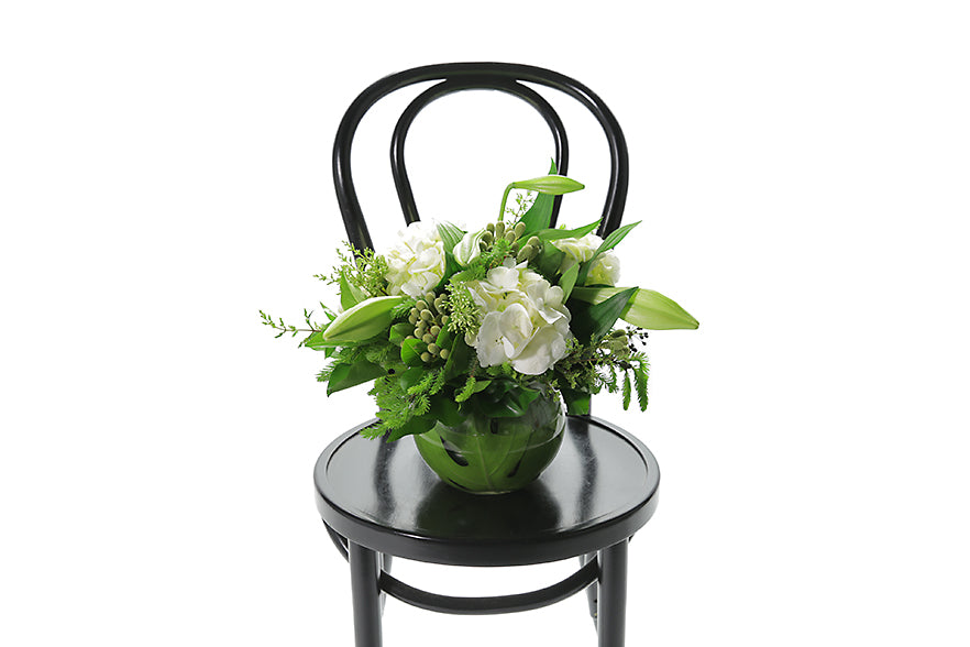 NOEL festive vase design is a classic vase design that displays a balanced mix of white seasonal festive flowers and green festive foliages. A ball vase lined with green monsteria leaves, displaying festive foliages, berries, christmas lilies and seasonal white and green flower. Ball vase sitting on a black bentwood chair.