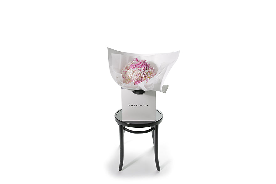 Wide image of the pink hydrangea bouquet. Small chic bouquet displaying a dome of pink hydrangeas (no additional foliage). Bouquet presented in Kate Hill Flower Bag. Bouquet bag sitting on black bentwood chair with white background.