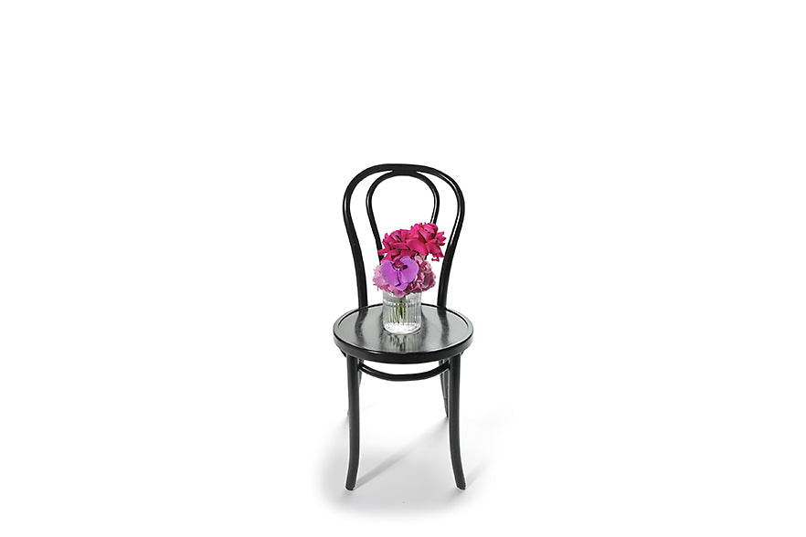 Wide image of the Royce vase design. Petite ribbed glass vase displaying bright pink and magenta seasonal flower. Gift includes the vase. Petite vase design sitting on black bentwood chair with white background.