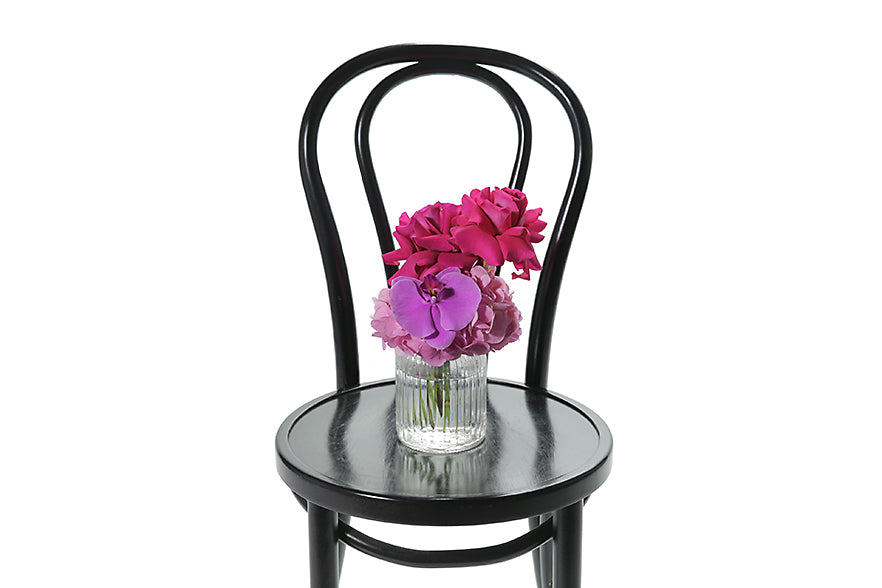 Petite ribbed glass vase displaying bright pink and magenta seasonal flower. Gift includes the vase. Petite vase design sitting on black bentwood chair with white background.