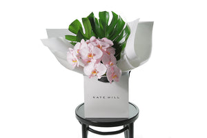 Chic and long lasting gift bouquet featuring 2 stems of blush phalaenopsis orchids and lush green monsteria leaves. Bouquet presented in Kate Hill Flower Bag. Orchid bouquet bag sitting on black bentwood chair with white background.