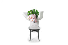 Wide image of the blush SADIE orchid flower bouquet. Chic and long lasting gift bouquet featuring 2 stems of blush phalaenopsis orchids and lush green monsteria leaves. Bouquet presented in Kate Hill Flower Bag. Orchid bouquet bag sitting on black bentwood chair with white background.