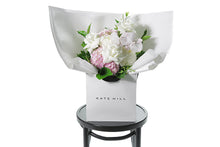 Pastel pink and white flower bouquet with seasonal foliage. Gift Bouquet presented in Kate Hill Flower Bag. Medium sized bouquet sitting on a black bentwood chair with white background.