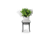Wide image of the VALERIE lily bouquet. Large gift bouquet displaying white oriental lilies (in bud not open) and green seasonal foliage. Gift bouquet presented in Kate Hill Flower Bag. Bouquet bag sitting on a black bentwood chair.