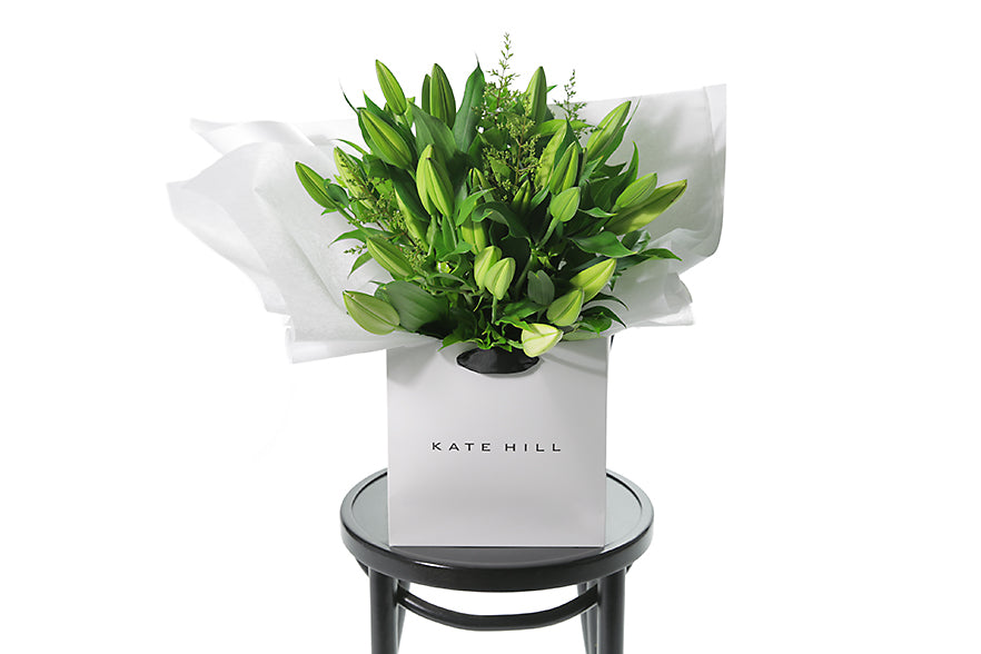 Large gift bouquet displaying white oriental lilies (in bud not open) and green seasonal foliage. Gift bouquet presented in Kate Hill Flower Bag. Bouquet bag sitting on a black bentwood chair.