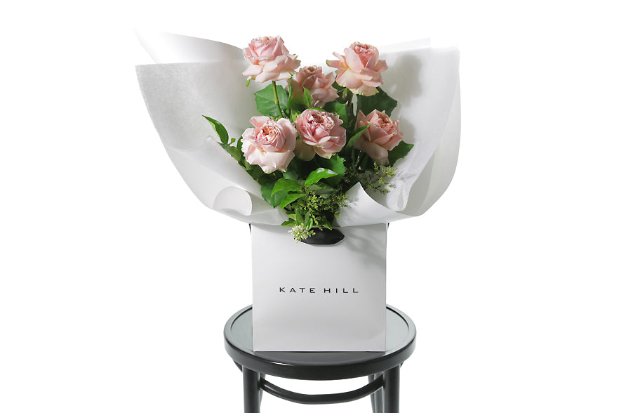 Classic rose bouquet featuring 6 stems of classic pink roses and green seasonal foliage. Gift bouquet presented in Kate Hill Flower Bag. Bouquet bag sitting on a black bentwood chair.