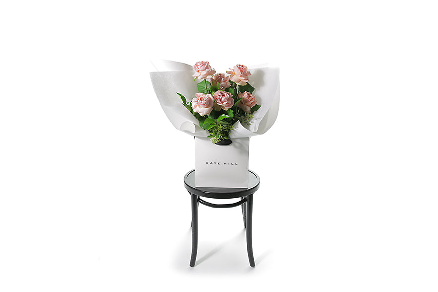 Wide image of the rose bouquet so you can see the size of the bouquet. Classic rose bouquet featuring 6 stems of classic pink roses and green seasonal foliage. Gift bouquet presented in Kate Hill Flower Bag. Bouquet bag sitting on a black bentwood chair.