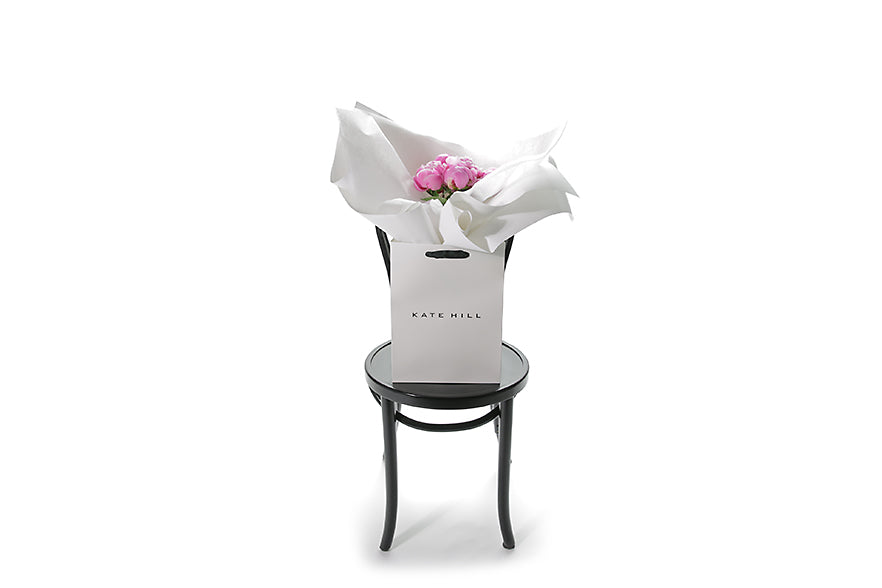 Wide image of the ZOEY peony rose bouquet. 5 Stems of seasonal pink peony roses gift wrapped beautifully in signature white wrapping. Bouquet presented in Kate Hill Flower Bag. Bouquet bag sitting on a black bentwood chair.