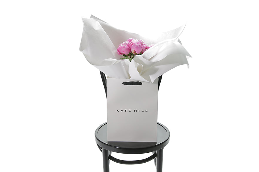 5 Stems of seasonal pink peony roses gift wrapped beautifully in signature white wrapping. Bouquet presented in Kate Hill Flower Bag. Bouquet bag sitting on a black bentwood chair.